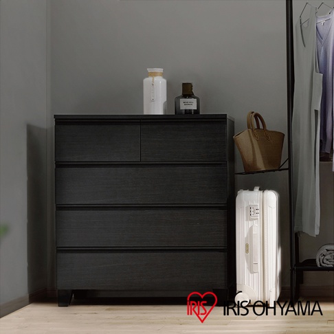 黑色木製收納櫃，有五個抽屜，底部有櫃腳，上方擺放著白色陶瓷瓶和香水瓶，旁邊有購物袋和白色行李箱。