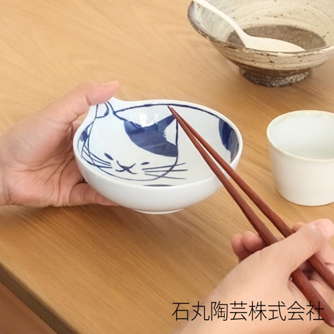 手持一只陶瓷貓咪湯碗，碗內有藍色貓咪臉部圖案，碗緣有貓耳朵造型。旁邊放有一雙木質筷子和一個白色小杯。