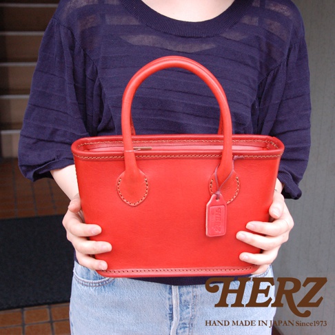 職人手作紅色皮革輕巧散步手提包，方形包身，配有圓形提把和同色系皮革吊牌，縫線清晰，展現皮革質感。