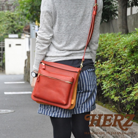 紅色皮革翻轉斜背包，側面為棕色皮革。包款為直式設計，設有拉鍊開口及前置拉鍊口袋。搭配細長皮革肩帶，由側面展示。