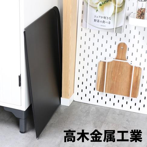 黑色 L 型折疊擋油板，可能為金屬材質，靠在白色櫥櫃旁，旁邊是木質立柱和白色洞洞板，掛有木質砧板。