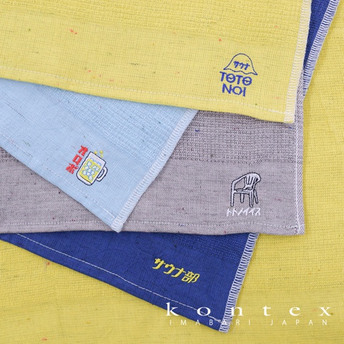 四條SAUNNER MOKU薄輕長精繡桑拿毛巾，包括黃色、淺藍色、灰色和藍色。黃色毛巾上有藍色「サウナ Tete NOI」字樣，淺藍色毛巾上有日文「フロ」和啤酒杯圖案，灰色毛巾上有椅子圖案及日文「トノイイス」字樣，藍色毛巾上有日文「サウナ部」字樣。毛巾材質為棉，表面有細微紋理。