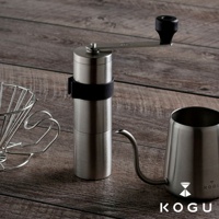 銀色18-8不鏽鋼手搖咖啡磨豆器，配有黑色手柄及黑色固定帶，置於木紋背景上，旁邊有同色系的金屬濾網架和細嘴咖啡壺。