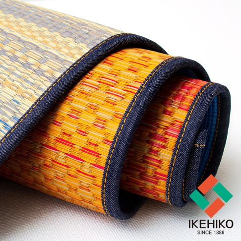 藺草編織的瑜珈墊，部分捲起，露出黃色、紅色交織的編織紋理，邊緣有深藍色滾邊及黃色縫線。