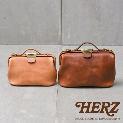 兩款日本HERZ職人手作皮革Organ鎖孔式大開口肩背包，一深棕色一淺棕色，並排陳列於灰色水泥背景前，下方有HERZ品牌標誌。
