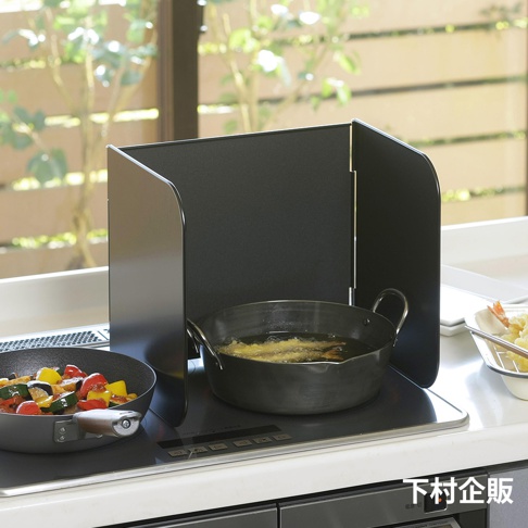 ㄇ型折疊防汙擋油板，黑色金屬材質，兩側擋板向內彎曲，放置於瓦斯爐上，中間有炒菜鍋和油炸鍋。