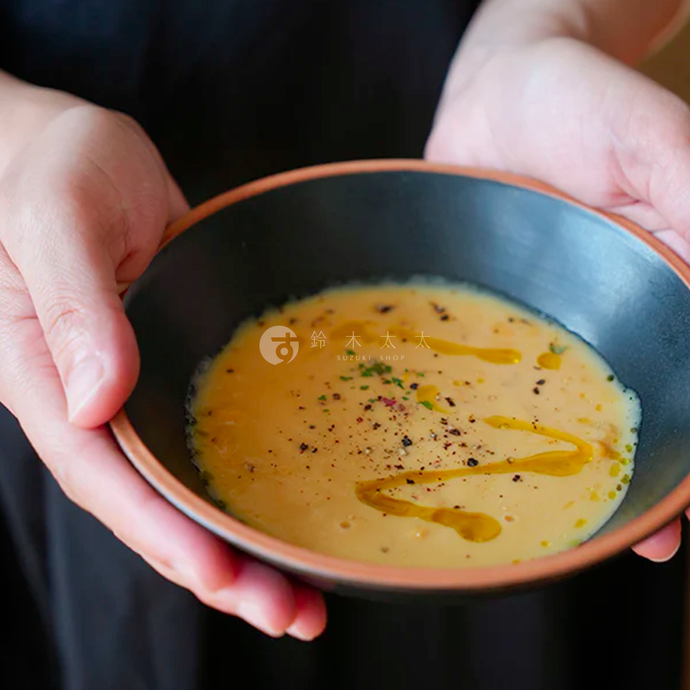 一只手捧著一個深色陶瓷沙拉碗，碗內盛有濃湯，湯面淋有橄欖油並撒有黑胡椒粒。