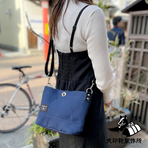 藍色燈芯絨迷你側背包，有細緻的條紋紋理，正面有一個銀色按扣和一個品牌標籤。背包透過黑色背帶斜挎在身。