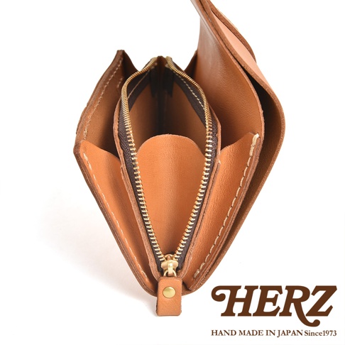由日本職人品牌 HERZ 手作的經典皮革皮夾，以俯視角度呈現，展現其棕色牛皮質感與細緻的縫線工藝，皮夾中間設有配備金屬拉鍊的零錢袋，右下方印有深棕色 HERZ 品牌標誌與 HAND MADE IN JAPAN Since1973 字樣。