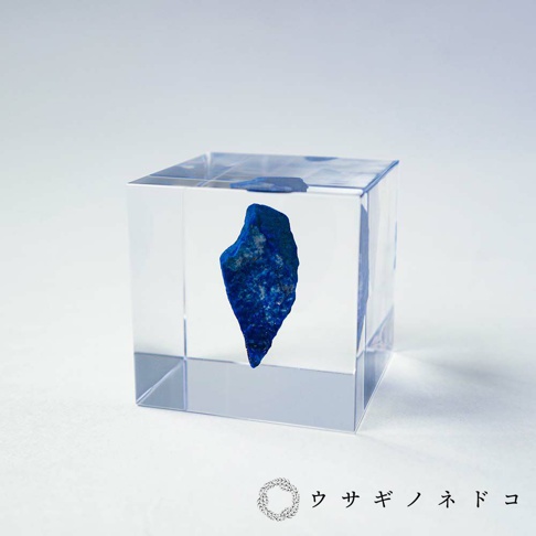 透明立方體中封存著一塊深藍色、帶有紋理的青金石礦石，礦石呈現不規則的塊狀，置於立方體中央，背景為白色。