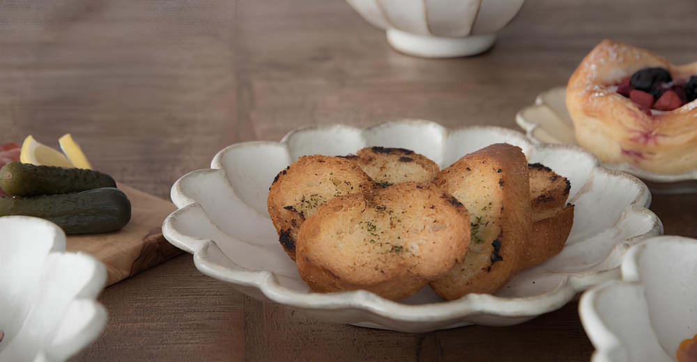 一個白色、帶有花瓣狀邊緣的陶瓷主食盤，盤中盛放著烤蒜香麵包片。