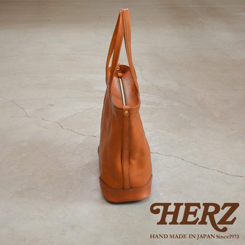 一個棕色皮革托特包，有拉鍊開合和兩個肩帶，擺放在水泥地上。右下方有 HERZ 的品牌標誌和 HAND MADE IN JAPAN Since1973 的字樣。