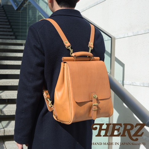 一位身穿深藍色外套的人背著一個棕褐色皮革後背包，背包設計風格類似屋瓦，配有可調整的皮革肩帶和金屬扣環。