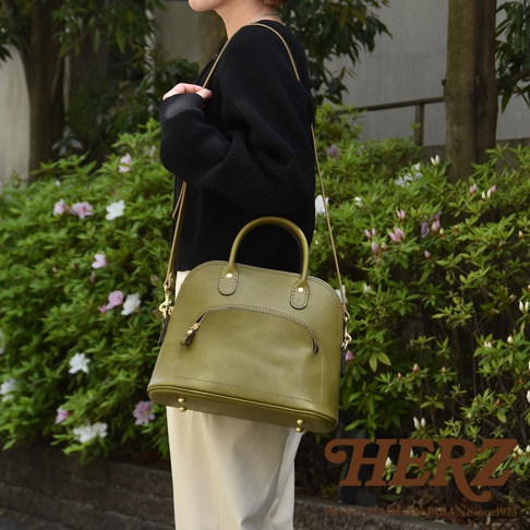 橄欖綠皮革兩用經典貝殼手提包，設有圓形手柄、可調節斜背帶及前方拉鍊口袋。包包底部有四個金屬腳釘。