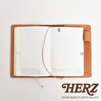 HERZ 品牌標誌，下方為 HAND MADE IN JAPAN Since1973 字樣，圖片中央為一本打開的棕色皮革手帳，內頁為日曆格式，以俯視角度拍攝。