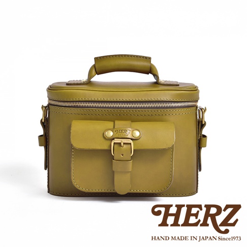 一手提皮革相機包，主體為橄欖綠色，有拉鍊封口，前方有一個帶金屬釦的翻蓋口袋，印有HERZ品牌標誌，背景為白色。