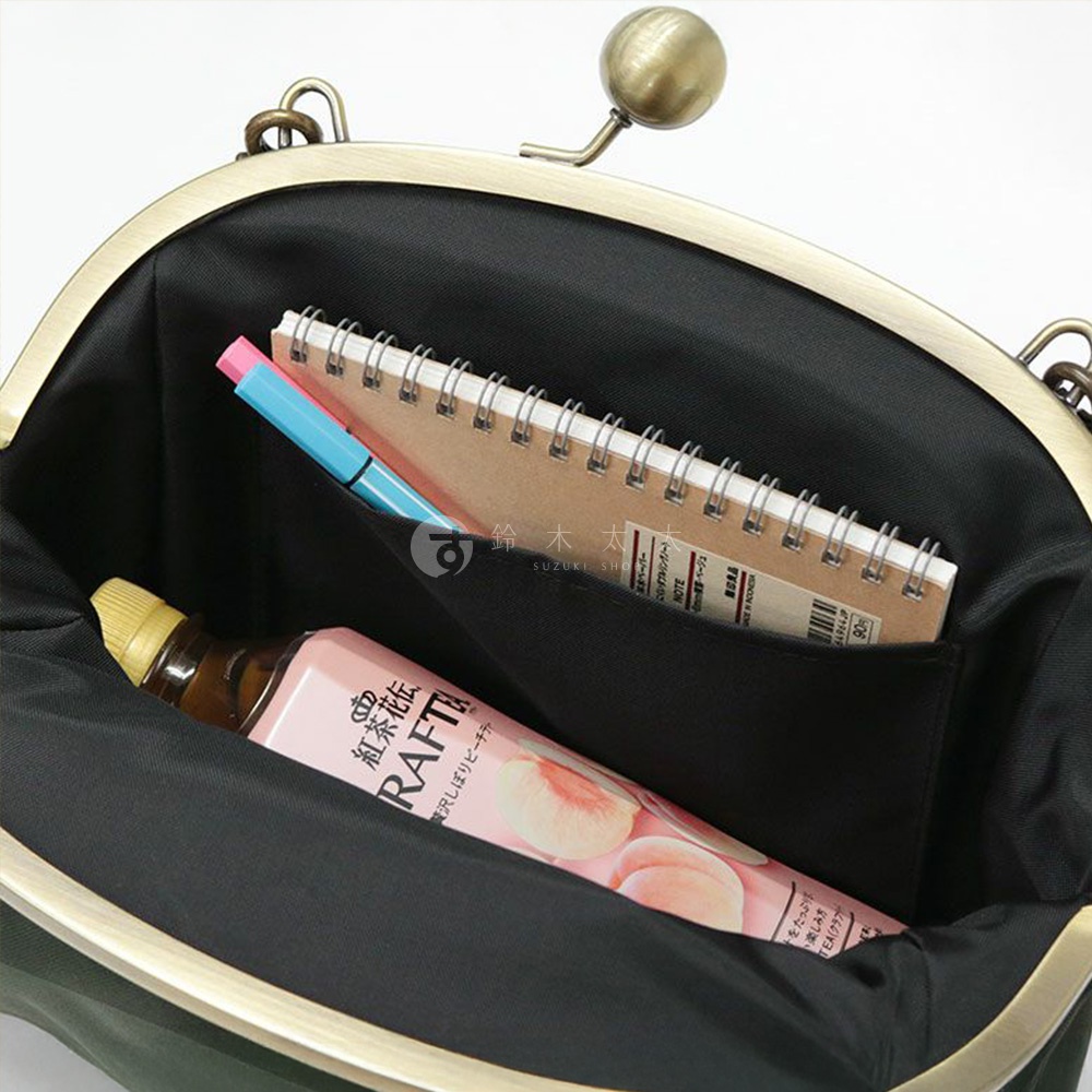 這款京都職人手作斜背包打開，內部有一個筆記本、一支粉色和藍色的筆，以及一瓶印有日文文字和水蜜桃圖案的飲料。