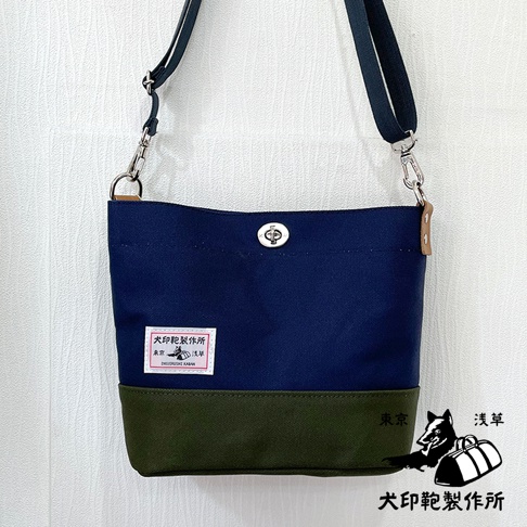 犬印純綿帆布雙色迷你側背包，主體為海軍藍，底部為軍綠色，配備可調節的深藍色織帶肩帶，銀色金屬扣具，正面有品牌標籤和金屬旋轉鎖扣。