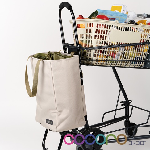 米白色帆布製手提收納袋，搭配軍綠色束繩內袋，懸掛於黑色摺疊推車側面。推車上層置有裝滿商品的購物籃。