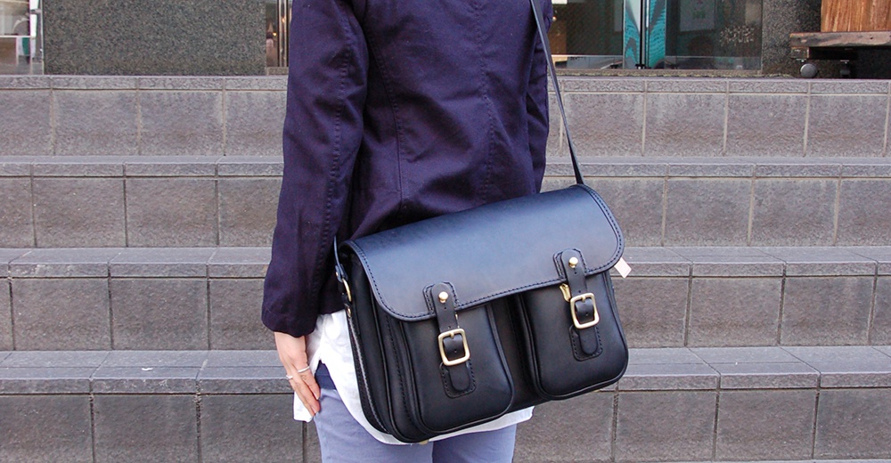 一位身穿深藍色外套、白色內搭和灰色長褲的人，肩背一個黑色皮革雙扣郵差包。