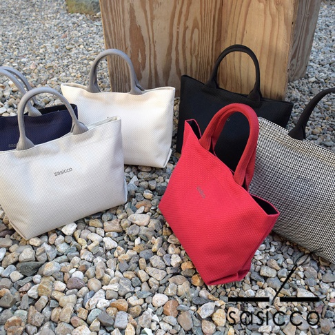 五個三河木綿刺子織托特包，顏色包含：淺灰色、白色、黑色、紅色及黑白格紋。包身帶有品牌 logo，手柄為灰色或黑色，皆置於碎石地面上。