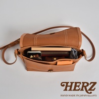 職人手作皮革復古郵差包，採用淺棕色皮革製作，包身線條簡潔。包內可見手機、錢包、書本等物品，整體呈現自然復古風格。