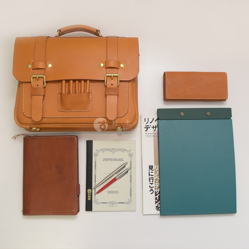 一咖棕褐色皮革製成、帶有傳統歐風設計的學生包，包包上有黃銅色金屬扣環和铆釘。包包旁擺放著同色系的長夾、兩本筆記本（一本為棕褐色皮革封面，另一本為帶有插畫的米白色封面）以及一支銀色和一支紅色筆，旁邊還有一本綠松石色的筆記本。