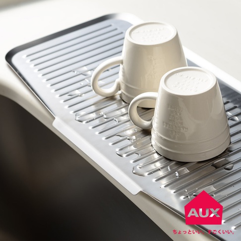 不鏽鋼長形瀝水籃上放置兩個白色馬克杯，籃身有條紋狀排水溝，右下角有品牌 Logo。