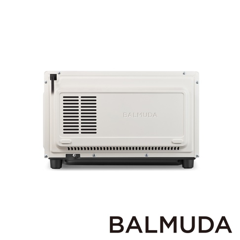 這是一張從正面角度拍攝的米白色 BALMUDA 烤箱背面特寫，金屬機身呈現簡約設計，左側設有垂直排列的散熱通風孔，底部邊緣有一排橫向小型散熱孔，下方裝有四個黑色支撐腳架，整體光線均勻，背景為純白色。