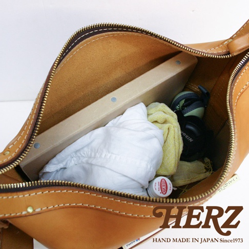 職人手作厚實皮革單肩波士頓包，展示內部結構，裝有白色衣物、黃色毛巾、相機及一瓶乳液。包包為棕褐色皮革，縫線清晰。