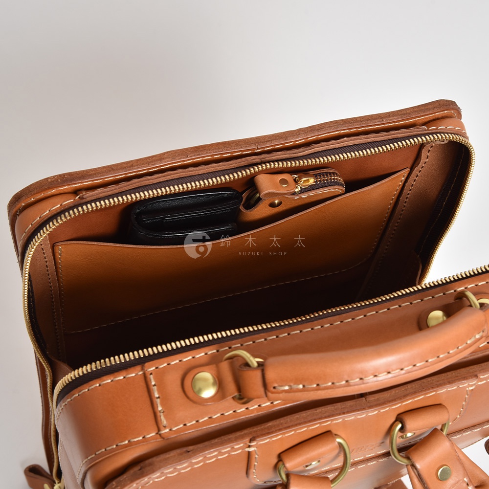職人手作皮革商務電腦後背包 R-25，展示了內部隔層，可見一個黑色皮夾放在卡其色皮革內袋中，背包以棕色皮革製成，搭配金色拉鍊和金屬配件。