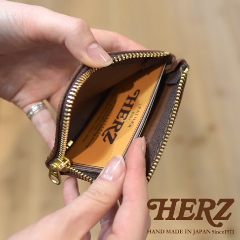 雙手正將一個棕色HERZ品牌皮革L型拉鍊收納夾打開，露出內部印有HERZ標誌的皮革卡片，整體呈現簡約精緻的日本職人手作風格。