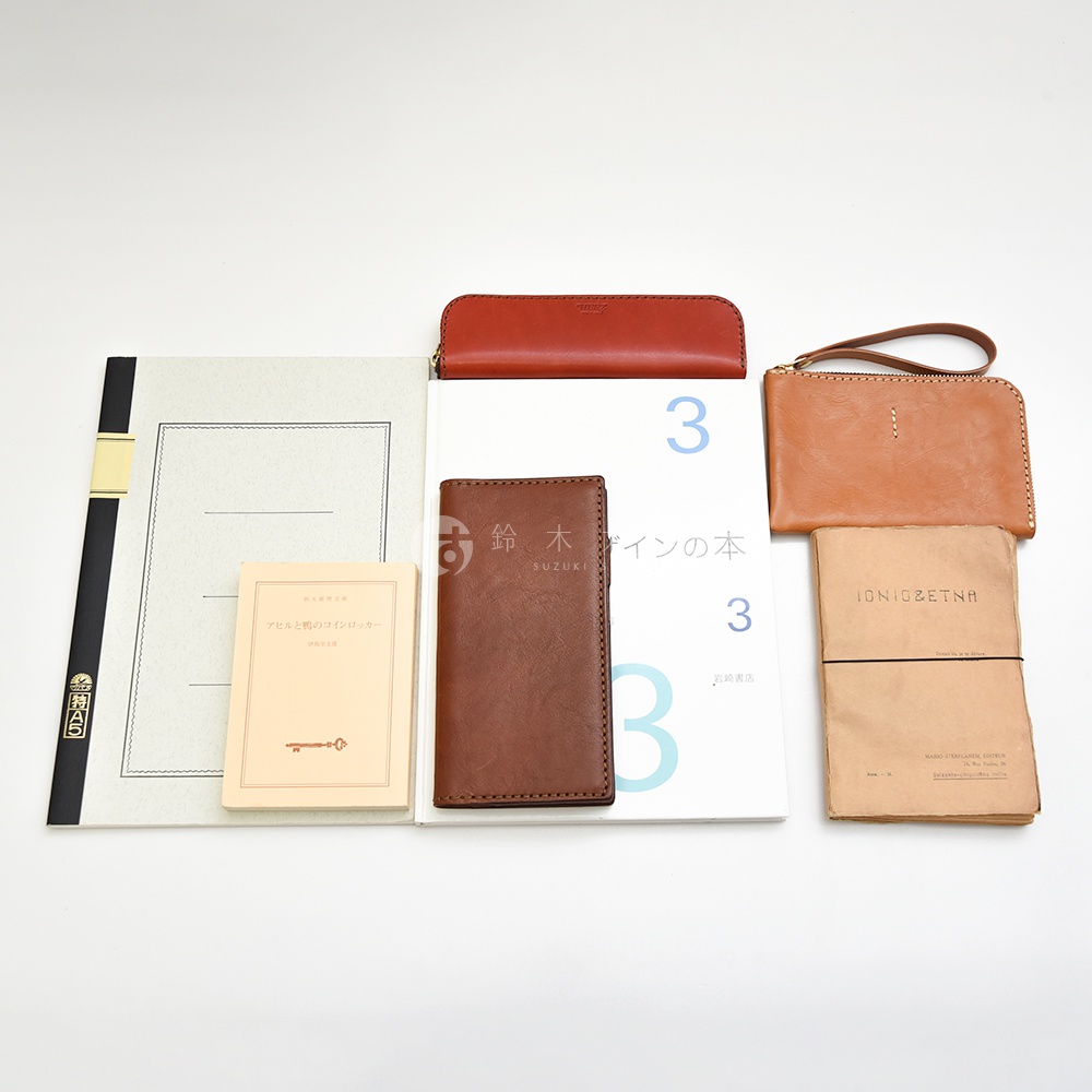 多款職人手作皮革製品，包含紅色長形筆夾、棕色長夾、淺棕色手拿包，以及兩本筆記本和一本小冊子。