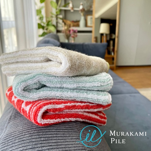 四條折疊的毛巾堆疊在灰色沙發上，最下面一條是紅白色條紋，中間是淺藍色，上面是米白色，左下角有 Murakami Pile 的藍色標誌。