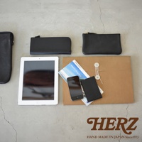 黑色皮革手拿包，包含一個長形拉鍊包、一個方形拉鍊包，以及一個較小的卡夾。旁邊擺放著一台平板電腦、一支手機、一本旅遊指南和一個牛皮紙封套。