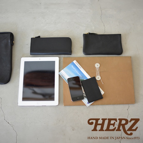 黑色皮革手拿包，包含一個長形拉鍊包、一個方形拉鍊包，以及一個較小的卡夾。旁邊擺放著一台平板電腦、一支手機、一本旅遊指南和一個牛皮紙封套。