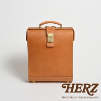 職人手作皮革杜勒斯商務後背包，淺棕色，配有金色鎖扣和提把，整體為方形設計，材質為皮革，底部有四個金屬腳釘。