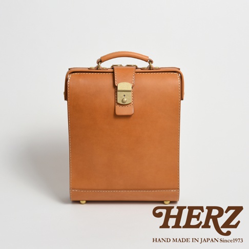 職人手作皮革杜勒斯商務後背包，淺棕色，配有金色鎖扣和提把，整體為方形設計，材質為皮革，底部有四個金屬腳釘。