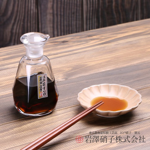 透明玻璃醬油瓶內裝有深褐色醬油，瓶身有日文標籤。旁邊擺放著一個淺色貝殼狀醬油碟，碟中盛有少量醬油，以及一雙木質筷子。