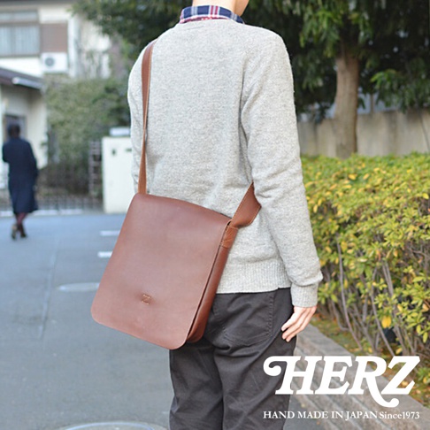 日本HERZ職人手作皮革軟皮直式斜背包，棕色皮革，以中景視角拍攝，搭配灰色毛衣和黑色褲子。