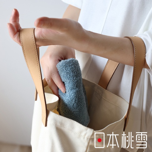 一隻手將一條淺藍色、柔軟的毛巾放入米白色帆布袋中，毛巾質地蓬鬆，袋子配有淺棕色皮革提把。