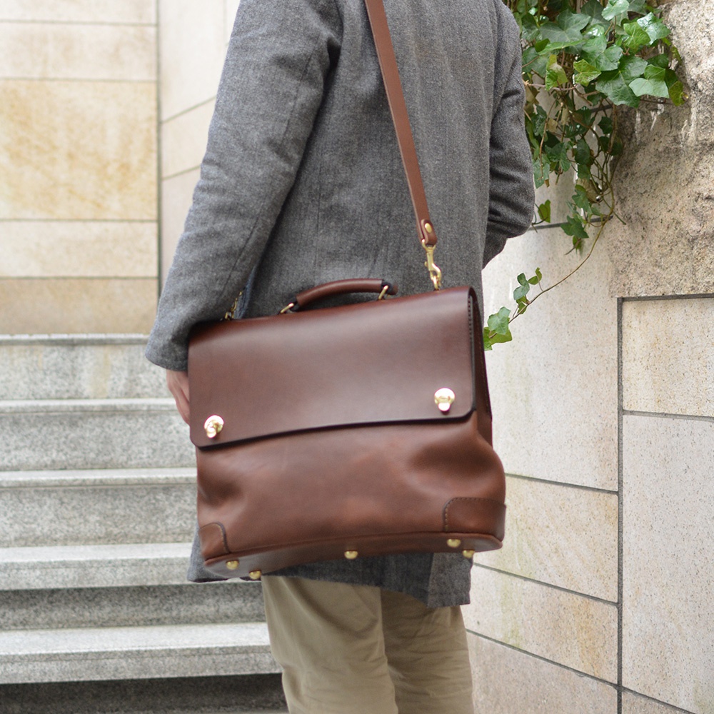 棕色皮革旅行商務兩用波士頓包，配有頂部提把、可拆卸肩帶和金色金屬扣。