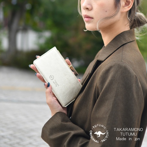 一手拿著一個淡金色壓紋皮革長夾，夾著一件棕色西裝外套，上面印有 TUTUMU 的標誌和 TAKARAMONO TUTUMU Made in jpn 字樣