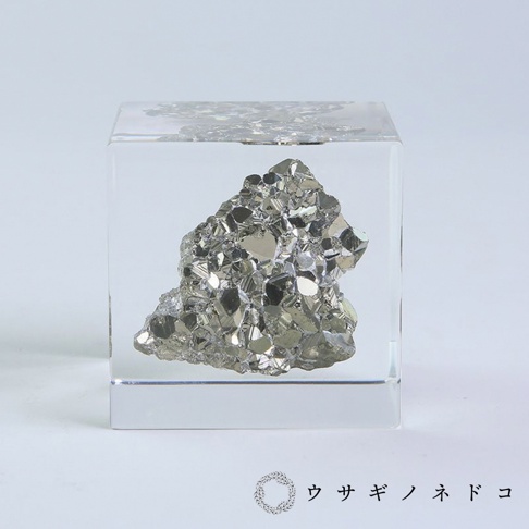 透明立方體中封存著一塊黃鐵礦，礦石呈現金屬光澤的銀灰色，表面有許多閃亮的結晶面，整體造型不規則，充滿立體感。