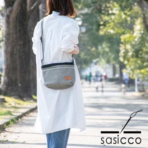 一位女性身穿白色長版上衣與牛仔褲，斜挎著一個黑色與白色細點圖案的三河木綿刺子織肩背包，包包上有一個淺棕色皮革標籤，揹帶為黑色鏈條。