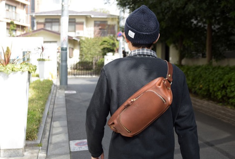 棕色皮革拉鍊旅行斜背包，斜挎於身穿深藍色毛衣的男子肩上，包身線條流暢，配有黃銅色拉鍊與金屬扣環，展現職人手作質感。
