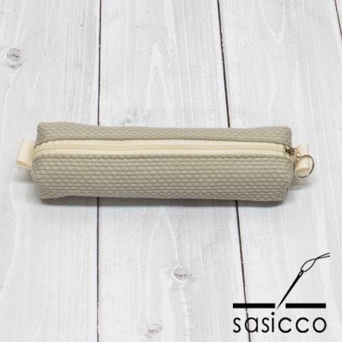 淺灰色三河木綿刺子織筆袋，配奶油色拉鍊及銀色拉環，左側有淺黃色織帶，置於淺色木紋背景上。