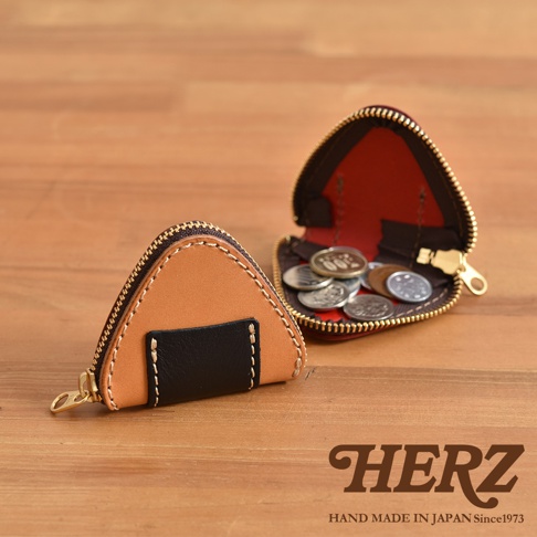 兩個日本HERZ手工皮革三角飯糰零錢包放置在木質桌面上，一個閉合展示外觀，另一個拉開拉鍊露出內部的硬幣，整體風格展現職人手工質感與細膩皮革工藝。