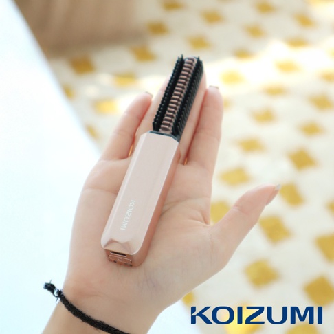 手持玫瑰金色迷你瀏海梳，梳齒為黑色，背景為模糊的黃色格紋布料。