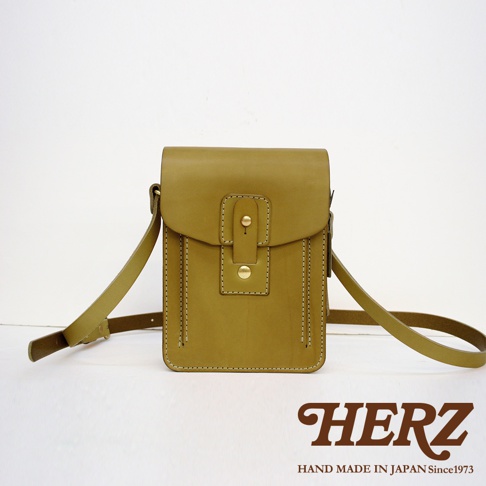 職人手作的淺棕色皮革斜背包，方形設計，正面有金屬釦環開合，搭配同色系皮革背帶，邊緣有縫線裝飾。